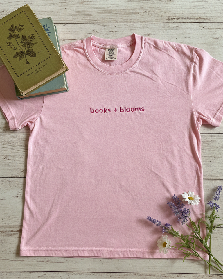 Pink t-shirt with 'books + blooms' text on a wooden surface with books and flowers.