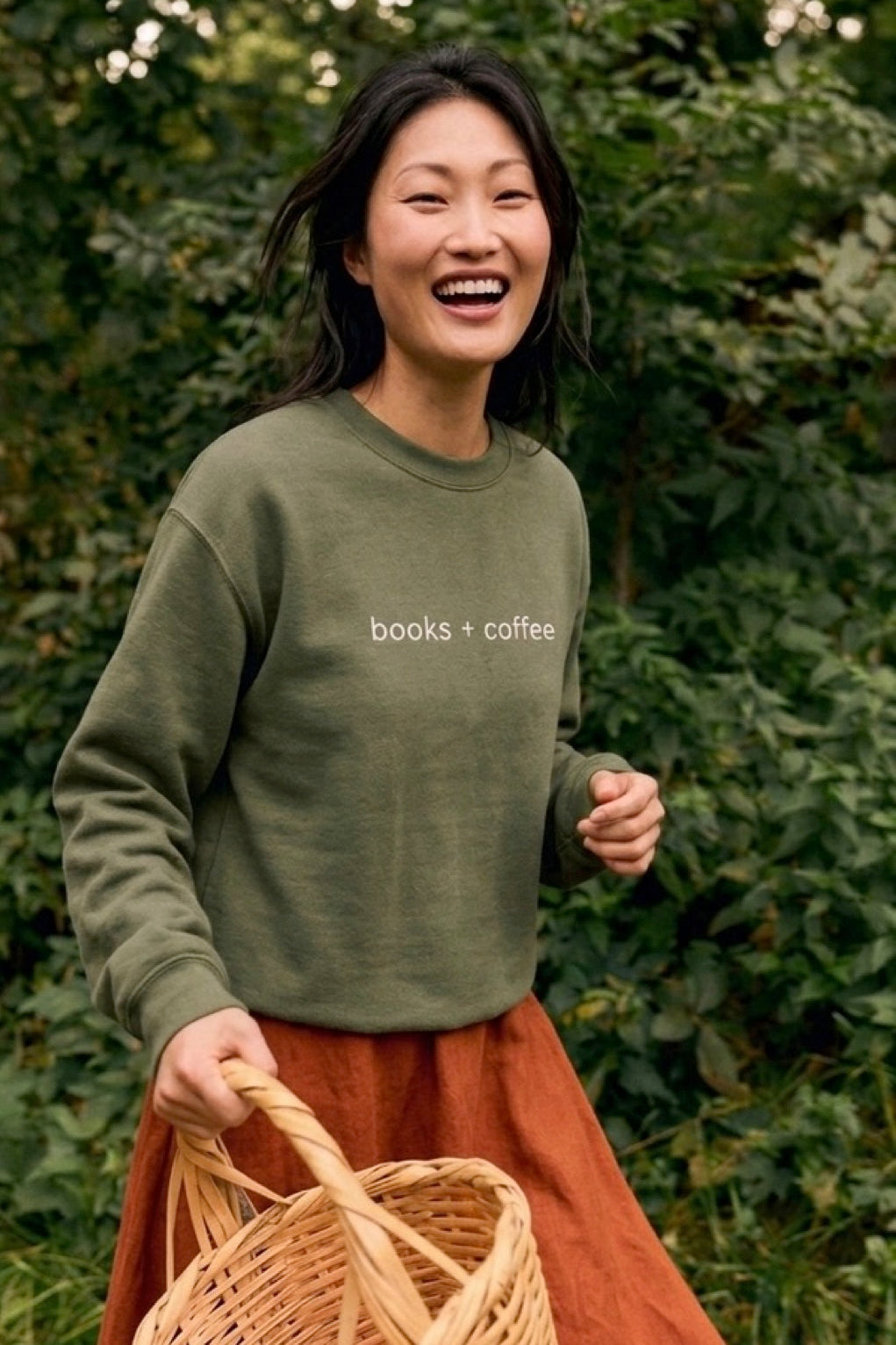 Person wearing a green sweatshirt with 'books + coffee' text, holding a woven basket, standing in front of green foliage.