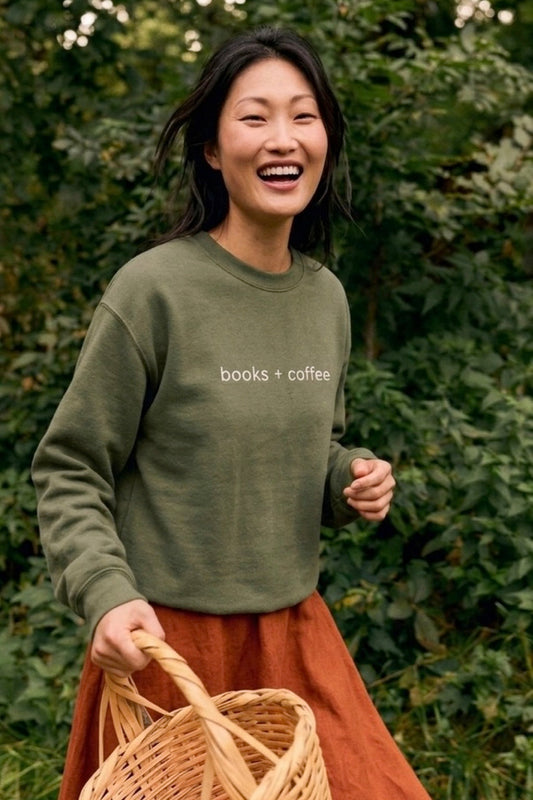 Person wearing a green sweatshirt with 'books + coffee' text, holding a woven basket, standing in front of green foliage.
